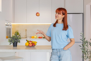 Young woman at home walks to bar counter picks apple from fruit bowl tosses it up takes bite smiles. Redhead girl in kitchen moves away happy step with fresh fruit snack positive mood morning vibe