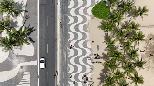 Top-down aerial of the Copacabana Mosaic Sidewalk in Rio de Janeiro, with its iconic black and white wave design, palm trees, beach sand, and busy road.