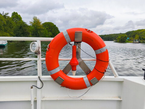 Berlin, Germany - September 05, 2025: Bright orange life buoy mounted on a boat railing, surrounded by tranquil water and lush green trees, emphasizing safety on the water
