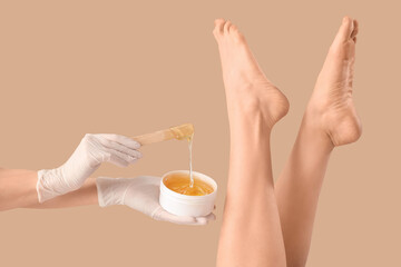 Beautician epilating legs of beautiful young woman with sugaring paste on brown background