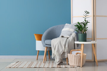 Armchair with cushion, plaid and houseplant on coffee table near blue wall in room