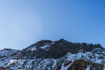 A rugged mountain ridge with scattered snow patches on dark rocks, framed against a clear blue sky