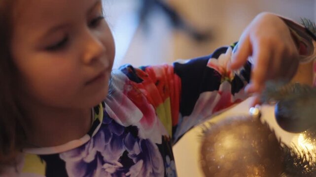 Little pretty girl dressing Christmas tree with large beautiful silver ball with sparkles at big family house. Close-up. Indoor. Winter time.