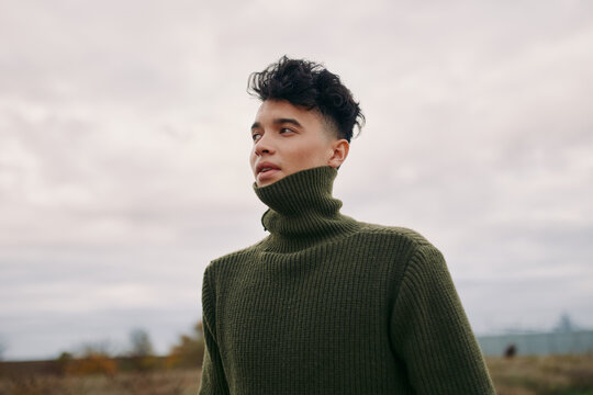 A young man in an olive knit turtleneck looks into the distance beneath a moody sky, a natural portrait emphasizing authenticity and credible documentary emotion with textured knit detail.
