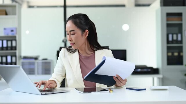 Asian woman using laptop and tablet while sitting at her working place. Concentrated at work.