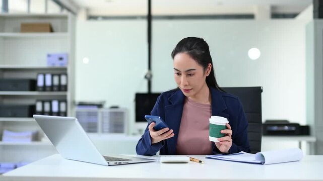 Asian woman relaxing with coffee and phone in modern office. Ideal for business, digital lifestyle, productivity, remote work, and workplace wellness stock footage.