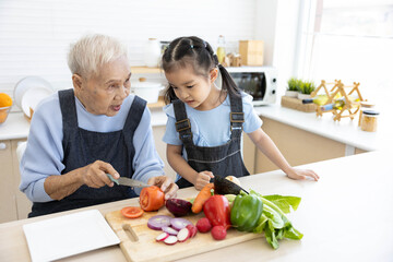 senior woman cutting fresh tomato and vegetables beside granddaughter waiting to eat in the kitchen