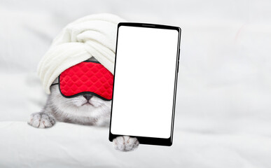 Cute kitten wearing sleeping mask and towel on it head lying on a bed at home and showing big...
