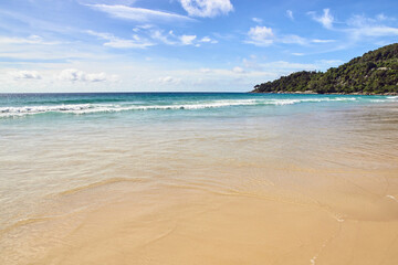 Tranquil sandy beach with clear blue waters and verdant green hills under a bright sunny sky.