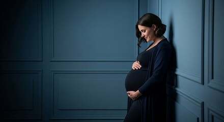 A pregnant woman gently holding her baby bump, studio portrait against a dark blue wall.