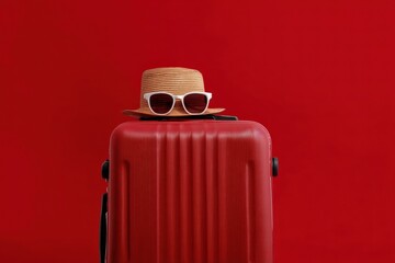 Red suitcase with sunglasses and straw hat on red background representing summer vacation and travel concept