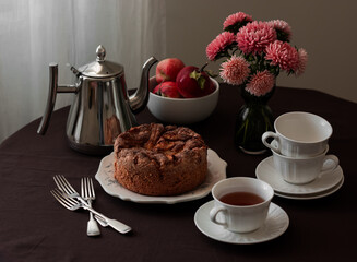 Homemade tea party - apple sponge cake, coffee pot, cups, flowers asters on a round table with a brown tablecloth