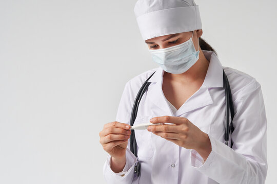 Doctor wearing face mask looking at measurement on medical thermometer over white background 