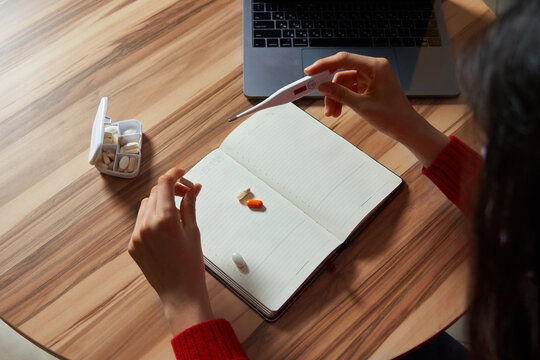 sick woman with pills and thermometer on a table working on a laptop from home on quarantine isolation.