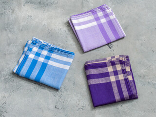 Three neatly folded, plaid men's handkerchiefs in shades of blue, purple, and lavender are arranged on a textured gray background.