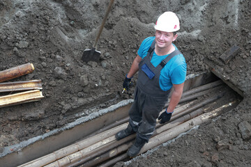 Caucasian male worker in construction gear standing in trench with pipes and soil.