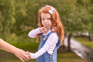 Obraz premium Red-haired caucasian young girl with blue bow in denim dress holding adult hand outdoors.