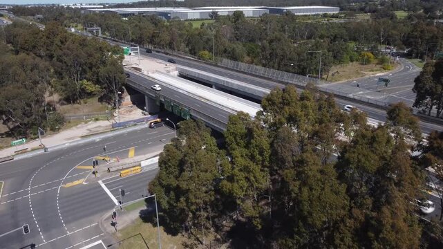 Backward drone shot over M7 motorway bridge with cars in motion and visible roadworks, elevated view of highway crossing secondary road in Western Sydney infrastructure scene