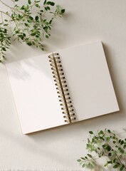 A blank spiral-bound notebook lies open on a light beige surface, adorned with sprigs of greenery on either side, casting soft shadows
