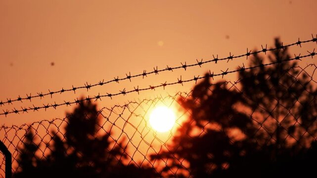 Dramatic sunset behind barbed wire fence showing freedom restriction, border security, imprisonment concept, orange sky silhouettes, cinematic mood, symbolic isolation visuals now