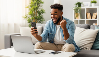 Excited African American man celebrating good news on his smartphone with a laptop nearby, depicting remote work success or online winning.

