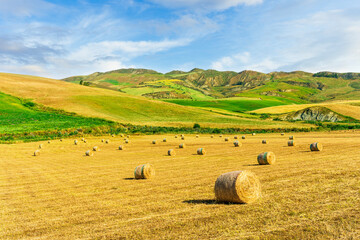 rustic landscape of a golden evening field with yellow wheat hay stacks among green hills and mountains and beautidul sunset cloudy sky on background. Summer season countryside landscape