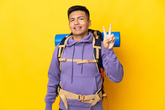 Young mountaineer man with a big backpack isolated on yellow background smiling and showing victory sign