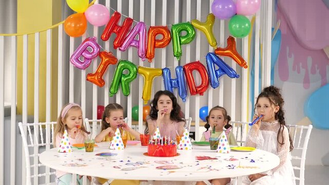 A group of smiling children in hats and with pipes at a birthday party. A HAPPY BIRTHDAY banner is displayed on the wall behind them.