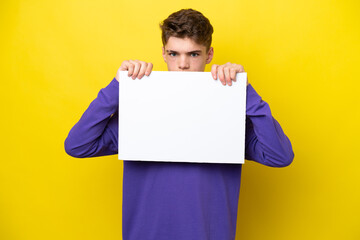 Teenager Russian man isolated on yellow background holding an empty placard and hiding behind it