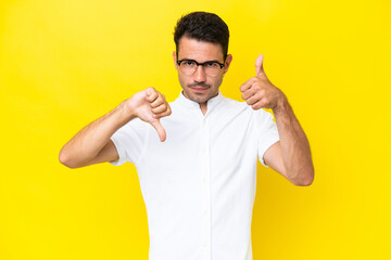 Young handsome man over isolated yellow background making good-bad sign. Undecided between yes or not