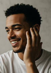 A smiling young man with curly hair in a white t-shirt, touching his face with one hand, looking away from the camera.