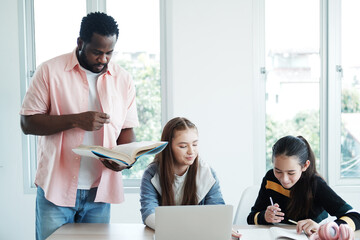 African American man is teacher or tutor teaching  and reading book for Caucasian students or pupils girl group in classroom at school. Education and technology concept
