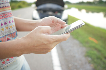 Close-up of a young woman's hand holding a smartphone to contact a repairman with a broken down car parked on the road in the background.