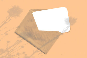 Natural light casts shadows from wildflowers on an envelope with a sheet of white paper lying on an orange background. Mockup