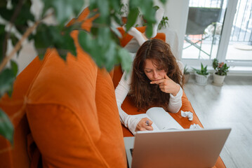 Pensive young woman lying on a comfortable orange sofa using laptop and taking notes on notebook having a creative crisis while working remotely from home