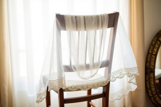 A delicate wedding veil with lace edges is draped over a wooden chair in a sunlit room. The background shows soft curtains and a vintage mirror