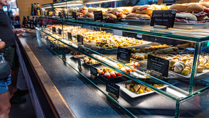 Freshly prepared pintxos featuring chicken skewers with curry-mango sauce and croquettes topped with padron peppers, displayed in a vibrant Ribera´s market stall in Bilbao.
