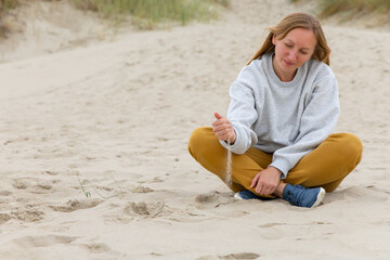Woman sitting cross-legged on sandy beach, gently letting sand slip through fingers, reflecting on nature and personal thoughts in serene environment