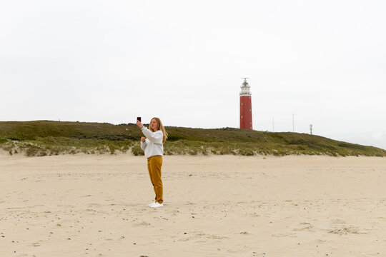 Individual standing on sandy beach, holding smartphone for video call, with lighthouse in the background, capturing distant connection concept