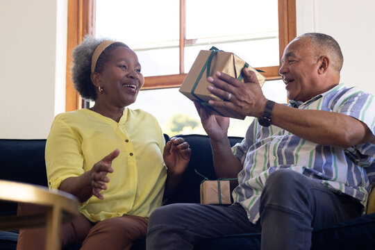 African American senior couple exchanging gift on couch, both looking joyful