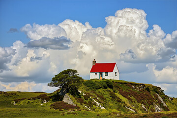 A solitary white cottage with a red roof on a green hill in Scotland. Idyllic rural landscape with a dramatic cloudy sky. Remote countryside living concept.