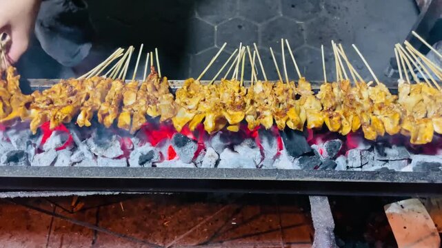 Street food vendor grilling chicken satay over traditional charcoal fire. The skewers are turned on an open flame, producing smoke and aroma in a lively street setting.