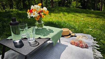Cozy summer picnic setup with flowers, drinks, fruit and a straw hat on a sunny lawn