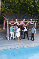 Exchanging hugs and flowers, diverse group celebrating by poolside under pergola