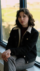 A young girl rests by a large window, gazing out thoughtfully. The scene highlights the blend of natural light, solitude, and youthful introspection.
