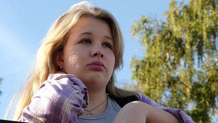 A blonde girl with tears in her eyes looks thoughtfully away, surrounded by a clear sky and trees. This image highlights themes of emotional depth and personal reflection.