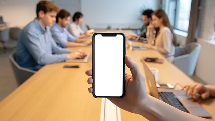 A person's hand holds a smartphone with a blank white screen, while a team of colleagues works in a blurred office meeting background.