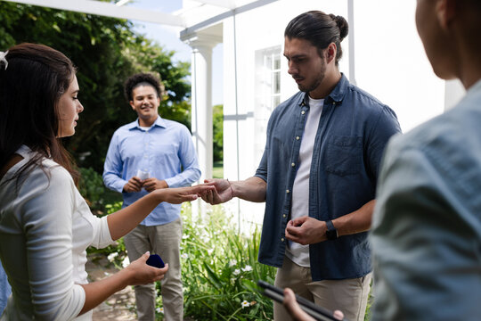 Exchanging rings outdoors, couple celebrating engagement with friends in garden