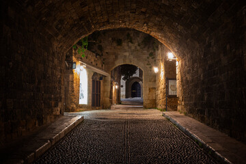 Fototapeta premium Idyllic and empty cobblestone street and tunnel at the medieval UNESCO World Heritage Site of Old Town of Rhodes on Rhodes island in Greece at night.