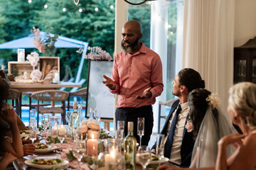 At wedding reception, man giving heartfelt speech, guests listening attentively around table